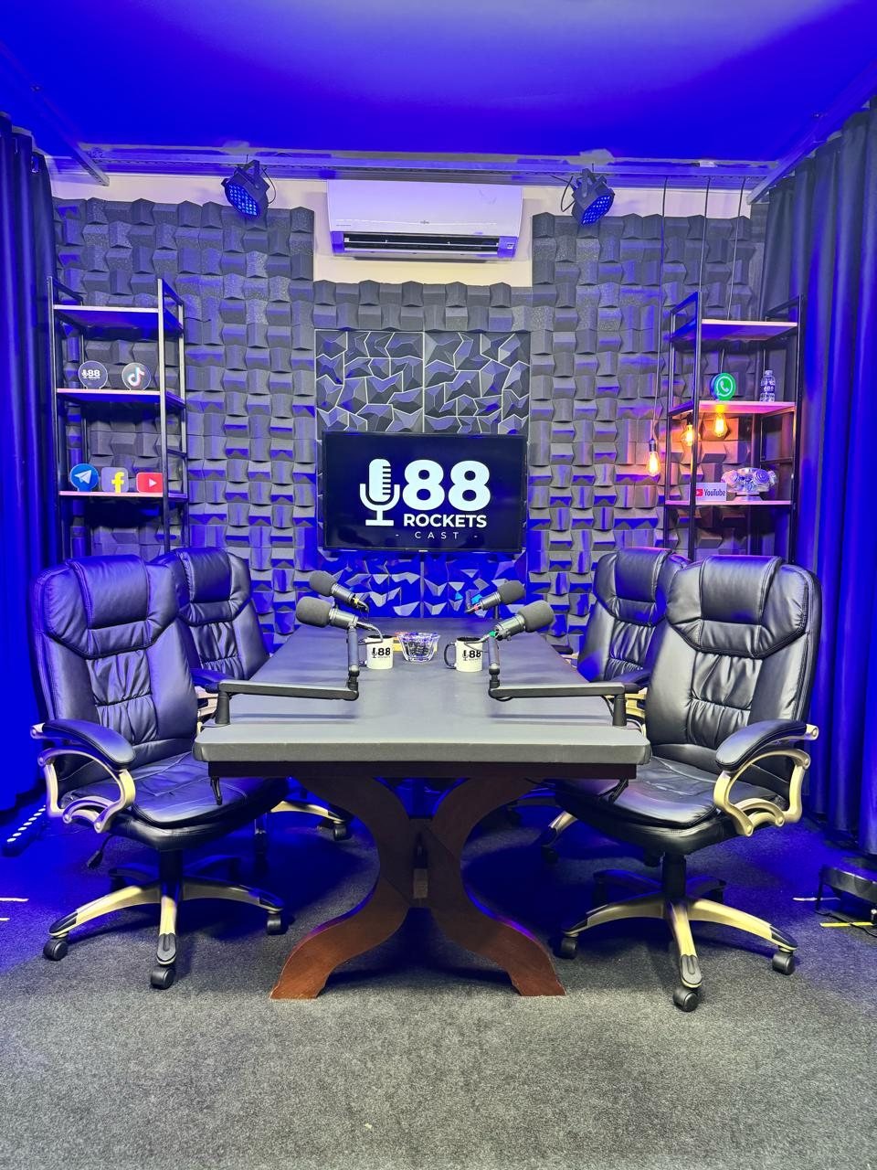 Estúdio de podcast em Campinas com mesa para quatro pessoas, microfones profissionais e parede acústica iluminada em azul